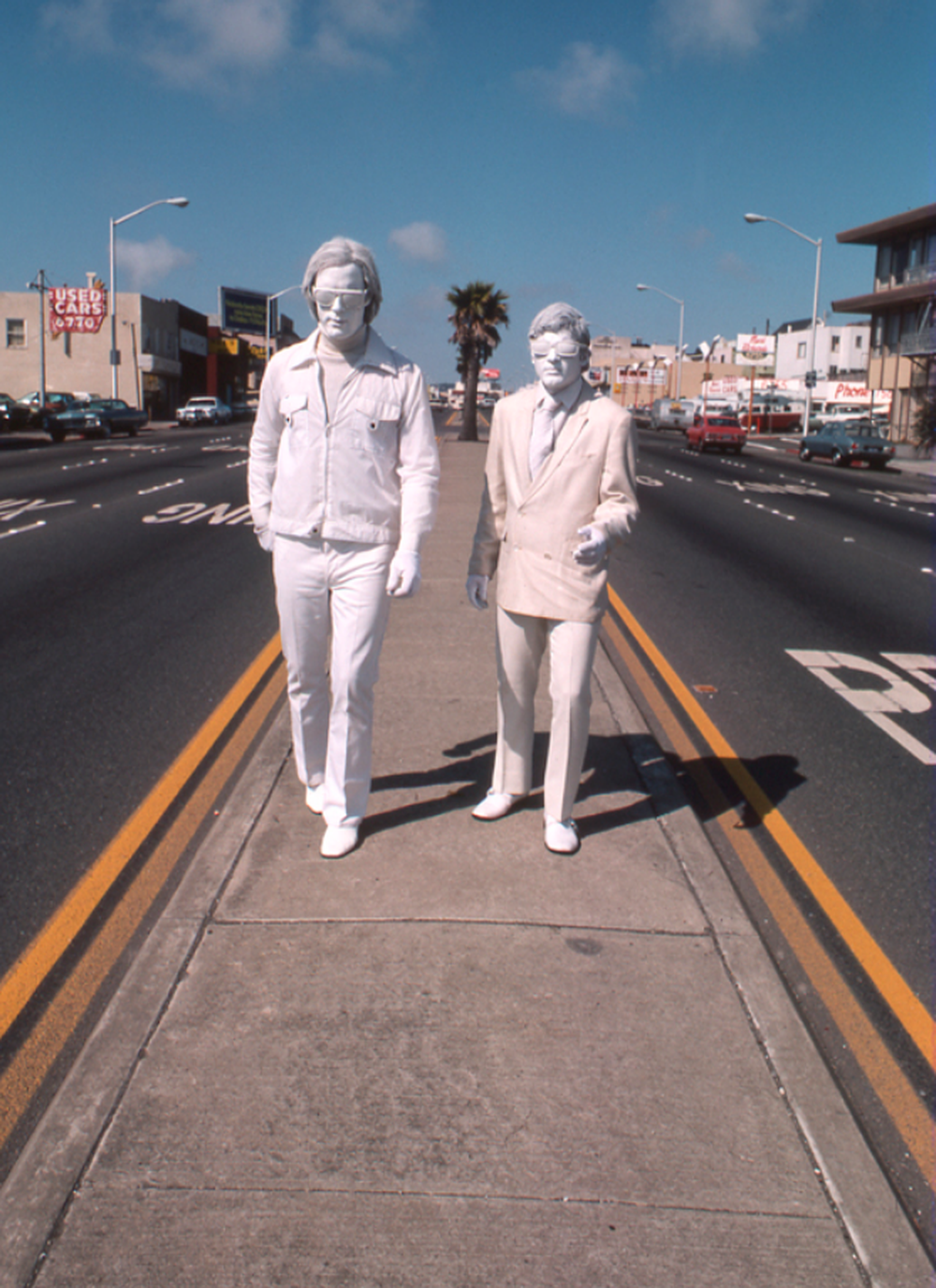 two men painted white wearing white clothing on the medium of a busy street