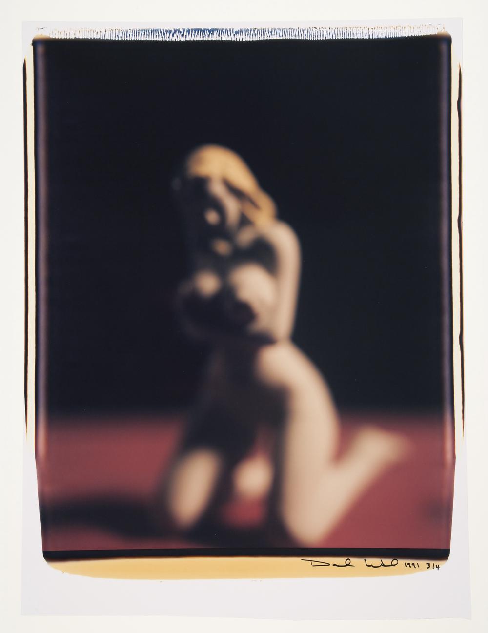 blurry image of nude blonde woman kneeling and facing the viewer while trying to cover herself