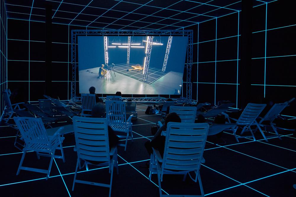 projection screen in a gallery with chairs for viewers in a darkened room with blue illumination
