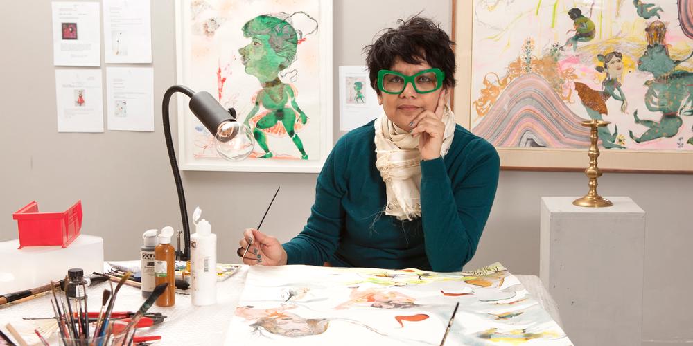 indian american woman seated at a desk with artwork on the wall behind her and a paintbrush in her hand. She is wearing large, bright, chunky green eyeglasses.