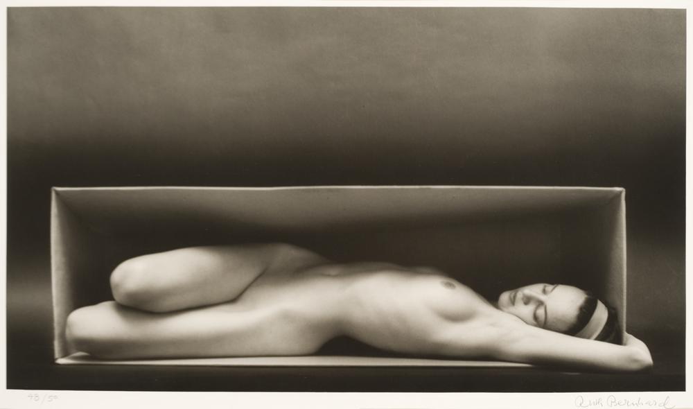 A nude woman laying in a horizontal box in a monochromatic background that centers the woman