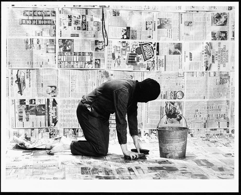 Man wearing a black shirt and pants and a ski mask in a wall papered with newspapers the man is holding two floor scrub brushed next to a metal bucket