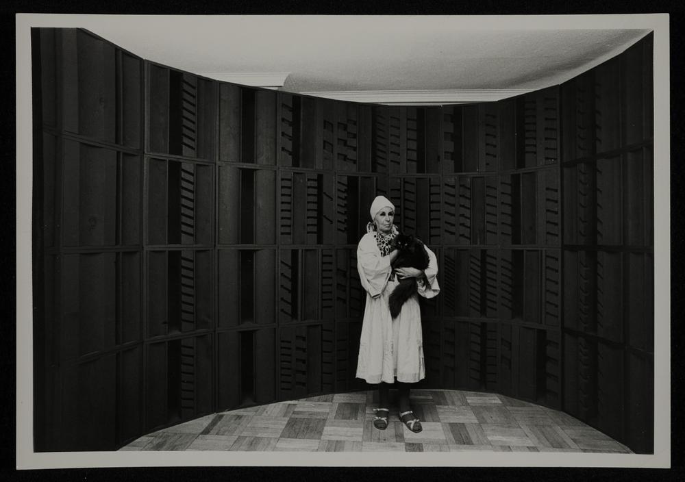 Nevelson dressed in white and standing in front of a wall sculpture with her black cat in her arms