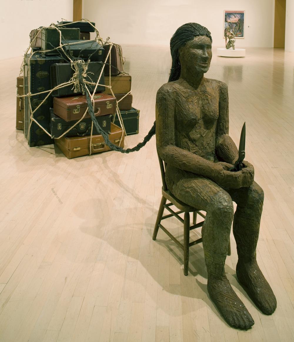 Installation view of a sculpted nude woman seated with her hair braided and attatched to suitcases that are tied into a bundle by rope